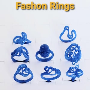 May include: A collection of nine blue fashion rings in various designs, displayed on individual white stands. The rings feature abstract and ornate shapes, with the text "Fashion Rings" in yellow above.