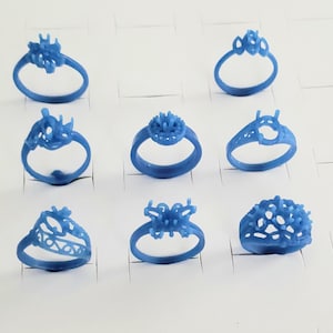 May include: Nine blue wax ring models in various designs, ready for the jewelry-making process. The rings feature different floral and geometric patterns, showcasing the intricate details of the designs. The rings are displayed on a white surface.