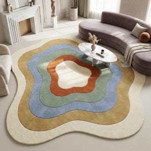Op de afbeelding: Een kleurrijk, abstract vloerkleed met een uniek, golvend ontwerp. Het kleed heeft concentrische lagen van crème, goud, blauw, groen en oranje. Het kleed is geplaatst in een woonkamer met een gebogen bank en een marmeren salontafel.