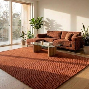 May include: A modern living room featuring a rust-colored rug with a ribbed texture, a matching sectional sofa, and a glass-top coffee table. Potted plants and a large window add natural elements to the space. The room has a warm, inviting atmosphere.