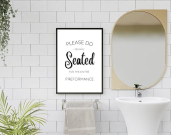 May include: A framed black and white sign with the text "PLEASE DO - REMAIN - Seated FOR THE ENTIRE PREFORMANCE" hangs on a white tiled wall. A gold-framed mirror and a white sink are also visible.