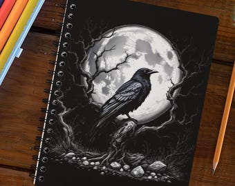 May include: A spiral-bound notebook featuring a black cover with a white illustration of a crow perched on a tree branch against a large full moon. Beside the notebook are two yellow pencils and a light blue plastic case containing markers.