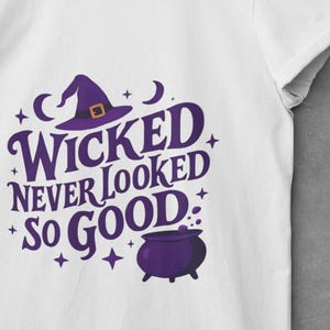 May include: White t-shirt with a purple graphic that reads "Wicked Never Looked So Good." The design includes a witch hat, cauldron, stars, and crescent moons. The shirt is displayed on a wooden hanger.