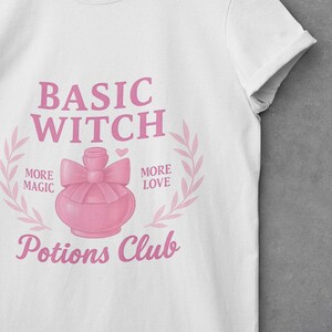 May include: White t-shirt featuring a pink graphic with the words "BASIC WITCH" and a pink potion bottle design. The shirt also includes the phrases "MORE MAGIC", "MORE LOVE", and "Potions Club" in pink.