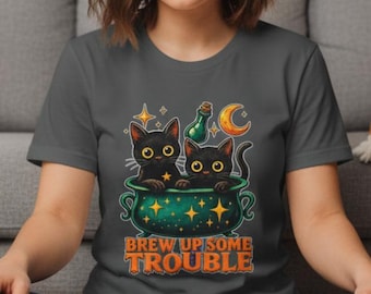 Op de afbeelding: Een antracietgrijs t-shirt met een afbeelding van twee zwarte katten in een groene ketel, met sterren, een halve maan en een flesje. De tekst "BREW UP SOME TROUBLE" staat onder de afbeelding in dikke oranje en gele letters.