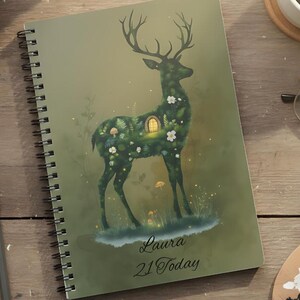 Può includere: Un taccuino a spirale verde con un'illustrazione di cervo piena di fiori e una casetta. Il testo "Laura 21 Today" è sotto il cervo. Il taccuino è su una superficie di legno con materiali artistici e una tazza di caffè.