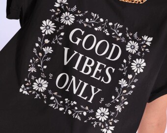 May include: Black t-shirt with the text "GOOD VIBES ONLY" in a white floral frame. The model is wearing tan pants and white sneakers. A red watch is on the model's wrist.
