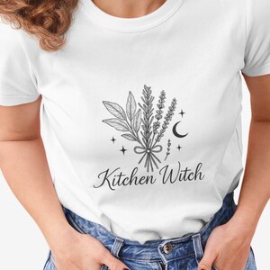 Könnte beinhalten: Weißes T-Shirt mit einer Schwarz-Weiß-Illustration von Kräutern, Sternen und einem Mond, die mit einem Band zusammengebunden sind. Die Worte "Kitchen Witch" stehen unter der Illustration in einer Schriftart.