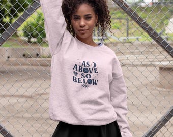 May include: A light pink sweatshirt with the text "AS ABOVE SO BELOW" in navy blue. The sweatshirt is paired with a black skirt. The image is taken outdoors with a chain-link fence in the background.