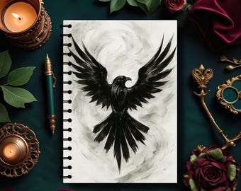 May include: A spiral-bound notebook featuring a black raven with outstretched wings against a smoky background. The image is surrounded by decorative elements, including a lit candle, a pen, and floral accents, all arranged on a dark green surface.