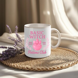 Puede incluir: Taza de cerámica blanca con el texto "BASIC WITCH" en rosa, junto con "MORE MAGIC" y "MORE LOVE". Un gráfico de botella de poción rosa está centrado debajo del texto, con "Potions Club" escrito debajo. La taza tiene un asa blanca y un borde dorado.