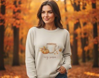 May include: A cream-colored sweatshirt featuring a watercolor design of a coffee mug, pumpkin, and fall leaves. The words "cozy season" are written below the design. The background is a blurred autumn scene with orange and yellow foliage.