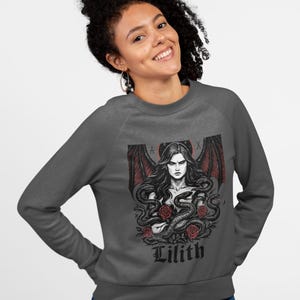 May include: Dark gray sweatshirt featuring a graphic of a woman with long hair, red bat wings, and snakes. The image includes red roses and the name "Lilith" in a gothic font. The sweatshirt is a crew neck style.