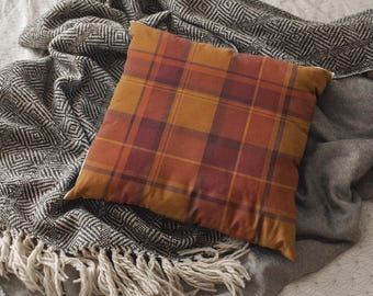 May include: A square pillow with a plaid pattern in shades of orange, red, and brown. The pillow is resting on a gray blanket with a geometric design and a fringed edge, and a solid gray blanket.