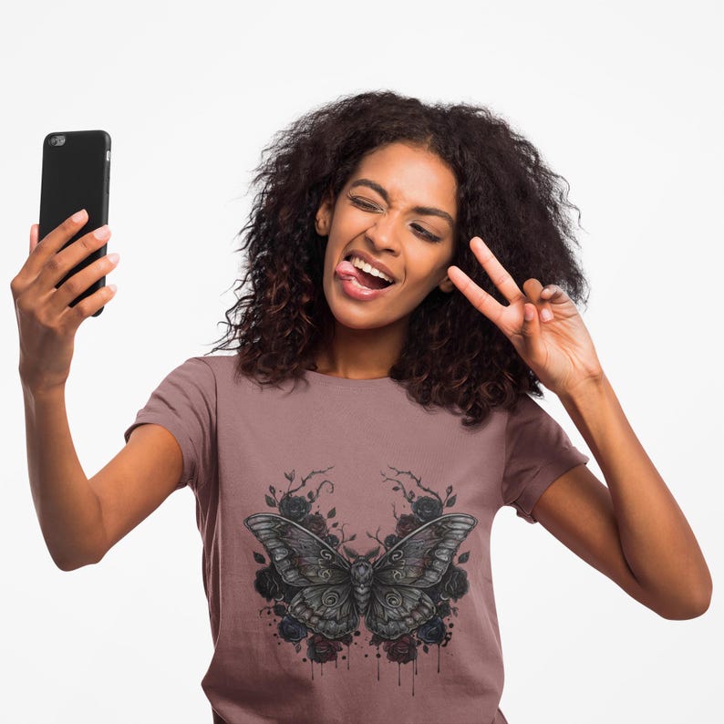 May include: A mauve t-shirt with a black and grey butterfly design and black roses. The person in the image is holding a black smartphone and making a peace sign with their fingers.