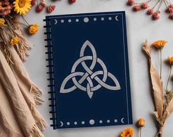 Puede incluir: Un cuaderno encuadernado en espiral con una cubierta azul marino con un diseño de nudo celta plateado. La cubierta también tiene un borde de lunas y puntos. El cuaderno está rodeado de flores y un paño beige.
