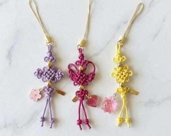 May include: Three decorative knotted cord charms with gold-tone loops. The left charm is lavender with a small pink flower, the center is magenta with a glittery heart, and the right is yellow with a translucent pink spherical bead.