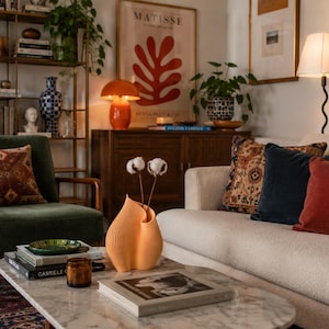 May include: A peach-coloured, ribbed, flame-shaped vase holding two dried cotton stems, positioned on a white marble coffee table. The vase features a vertical, undulating texture and a curved opening, serving as a central decorative object.