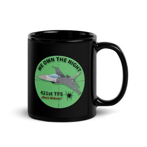 May include: Black ceramic mug with a green circular emblem. The emblem features a fighter jet in a crosshair, the text "WE OWN THE NIGHT", and "421st TFS Black Widows". The mug has a large handle.