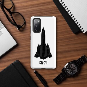 May include: A white phone case with a black SR-71 aircraft design and the text "SR-71". The case is on a wooden surface, with a laptop, glasses, a watch, a pen, and a notebook. The design is a detailed illustration of the iconic aircraft.