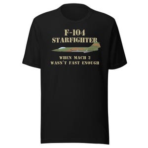 Puede incluir: Camiseta negra con el texto "F-104 STARFIGHTER WHEN MACH 2 WASN'T FAST ENOUGH" y una ilustración de un avión de combate verde y marrón. La camiseta está hecha de un material suave.