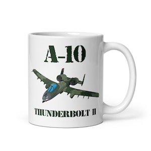 Puede incluir: Taza de cerámica blanca con una ilustración verde de un avión A-10 Thunderbolt II. La taza tiene el texto "A-10" encima del avión y "THUNDERBOLT II" debajo, todo en color verde oscuro.