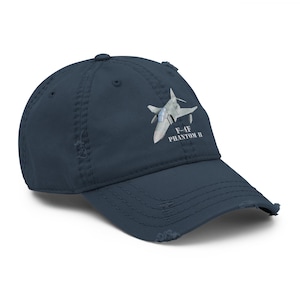 Peut inclure: Casquette de baseball bleu marine délavé avec une illustration d'un avion de chasse. Le texte "F-4E PHANTOM II" est imprimé en blanc. La casquette a un aspect usé avec des bords effilochés.