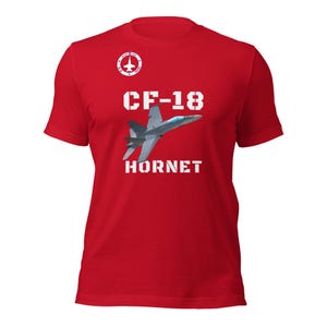 May include: Red t-shirt featuring a graphic of a CF-18 Hornet fighter jet. The design includes the text "CF-18" above the jet and "HORNET" below it, all in white block letters. A circular emblem with a jet is at the top left.