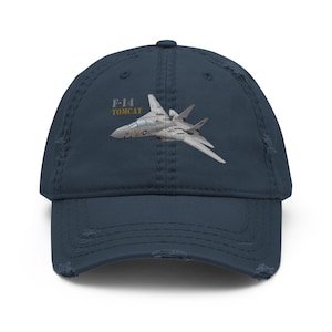 Peut inclure: Une casquette de baseball bleu marine à l'aspect usé, ornée d'une broderie représentant un avion de chasse F-14 Tomcat gris en vol. Le texte F-14 TOMCAT est brodé en gris et jaune au-dessus de l'appareil. La visière et les panneaux sont effilochés.