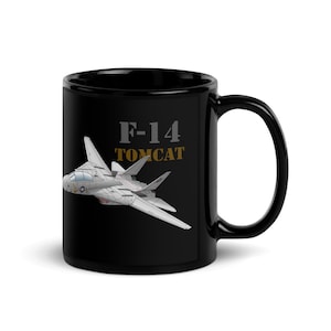 May include: A black ceramic mug featuring a detailed illustration of a grey F-14 Tomcat fighter jet in flight. The text F-14 is printed in grey above the aircraft, with the word TOMCAT printed in gold lettering directly below the model number.