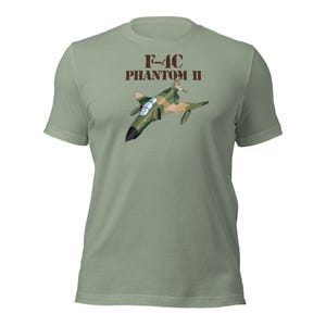 Può includere: T-shirt verde chiaro con grafica di un jet da combattimento F-4C Phantom II nei colori mimetici. Il testo "F-4C PHANTOM II" è stampato sopra il jet in marrone. La maglietta ha maniche corte e girocollo.