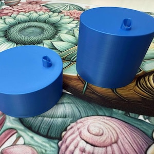 May include: Two cylindrical blue objects with a small loop on top. One is shorter and wider than the other. The objects are on a colorful floral and seashell patterned surface. The objects appear to be bottle stoppers.