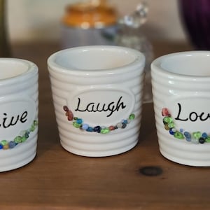 Op de afbeelding: Drie witte cilindervormige keramische houders met horizontale ribbels. Elke houder heeft een ovaal plaatje met de woorden Live, Laugh en Love in zwarte schrijfletters, geaccentueerd door een kleine halve cirkel van gekleurde glaskralen eronder.