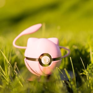 May include: A pink, spherical toy with a cat-like face, black accents, and a curved antenna. The toy is set in a grassy field, with a blurred green background.