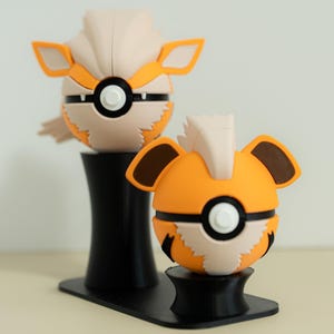 May include: Two orange and tan character figures, resembling a stylized animal, are displayed on a black stand. The figures have black and white accents, with a round shape and prominent ears. The stand has a curved design.