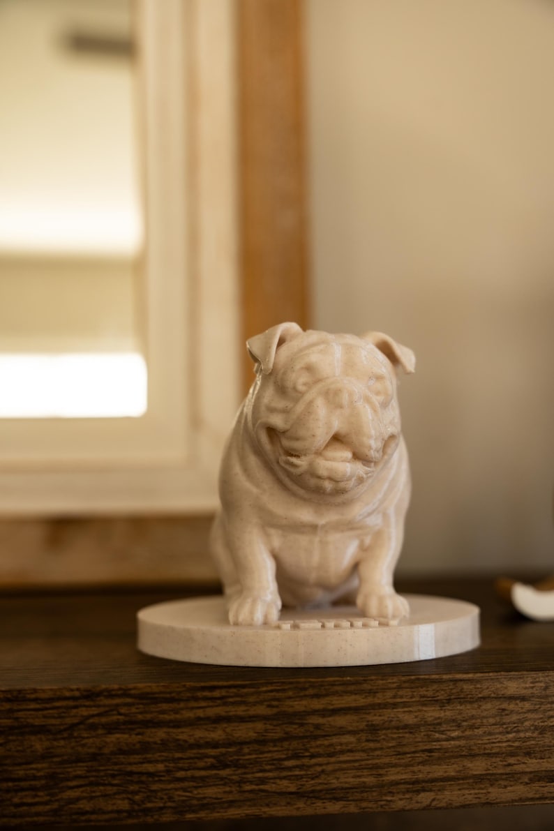 May include: A beige bulldog figurine sitting on a round base. The bulldog is sculpted with detailed features, including wrinkles and a slight smile. The figurine is made of a smooth, possibly stone-like material.