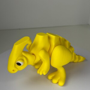 May include: A bright yellow, articulated dinosaur toy with a detailed design. The toy features a head, body segments, legs, and a tail. The dinosaur has a small eye and is posed in a walking position. The toy is made of plastic.