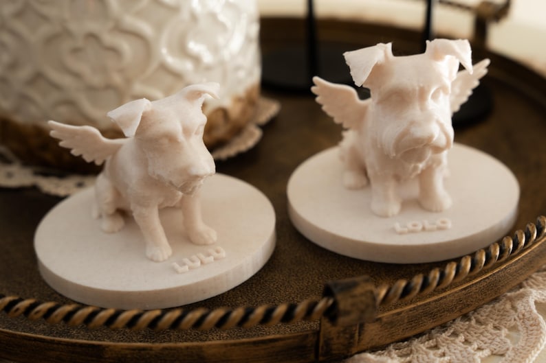 May include: Two off-white dog figurines with angel wings, each sitting on a round base. The name "LULU" is inscribed on the base of one figurine. The figurines are on a decorative tray.