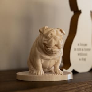 May include: A light brown wooden bulldog figurine sitting on a round base. The bulldog is detailed with wrinkles and folds, and is a decorative item. A wooden silhouette of a dog is in the background.