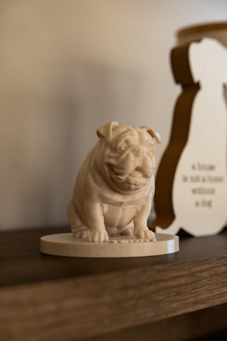 May include: A light brown wooden bulldog figurine sitting on a round base. The bulldog is detailed with wrinkles and folds, and is a decorative item. A wooden silhouette of a dog is in the background.