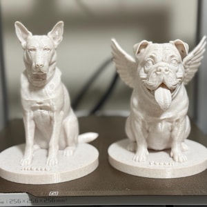 May include: Two off-white dog sculptures. One is a sitting Bull Terrier. The other is a Bulldog with angel wings and its tongue sticking out. Both are on round bases. The sculptures are likely 3D printed.