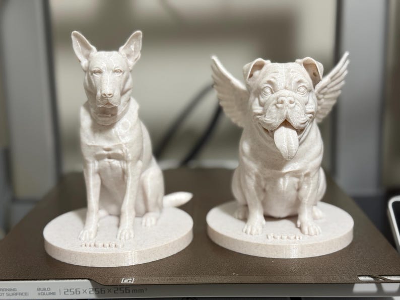May include: Two off-white dog sculptures. One is a sitting Bull Terrier. The other is a Bulldog with angel wings and its tongue sticking out. Both are on round bases. The sculptures are likely 3D printed.