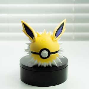 May include: A yellow and white Jolteon figure with blue accents, resembling a Poké Ball. The figure has spiky details and is set on a black circular base. The ears are yellow with blue tips.