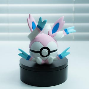 May include: A Sylveon figurine, a Pokémon character, is displayed on a black circular base. The figure features a pink and white body with blue and white accents. The figure has multiple ribbon-like appendages.