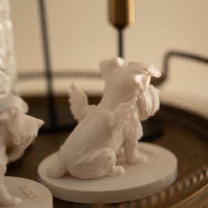 May include: A white figurine of a dog with angel wings, sitting on a round base. The dog has a fluffy face and tail. The figurine is on a decorative tray with gold candle holders in the background.