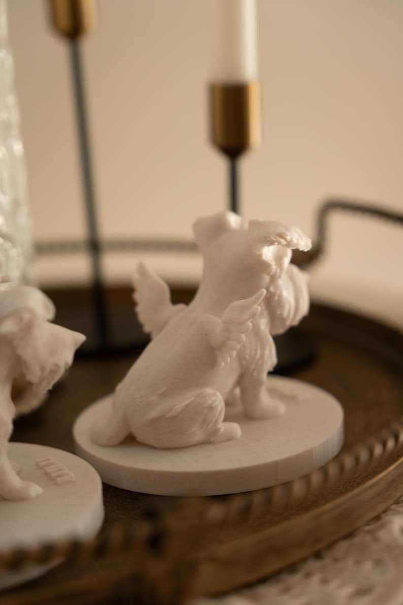 May include: A white figurine of a dog with angel wings, sitting on a round base. The dog has a fluffy face and tail. The figurine is on a decorative tray with gold candle holders in the background.