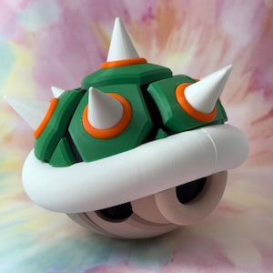 Peut inclure: Figurine de carapace de Bowser verte, blanche et orange. La carapace a des pointes blanches, des accents orange et un bord blanc. La partie inférieure est beige avec deux trous noirs pour les yeux.