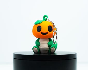 Könnte beinhalten: Ein Kürbis-Schlüsselanhänger-Figur mit einem orangefarbenen Kopf, einem grünen Stiel und einem grünen Körper. Die Figur hat ein lächelndes Gesicht und ist an einem silbernen Schlüsselanhänger befestigt. Der Artikel steht auf einer schwarzen Oberfläche.