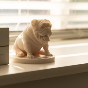May include: A light pink bulldog figurine with an open mouth, sitting on a round base. The figurine is on a white surface, with a wooden block with the words "kids paws" in the background.