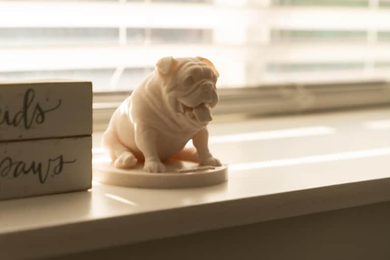 May include: A light pink bulldog figurine with an open mouth, sitting on a round base. The figurine is on a white surface, with a wooden block with the words "kids paws" in the background.